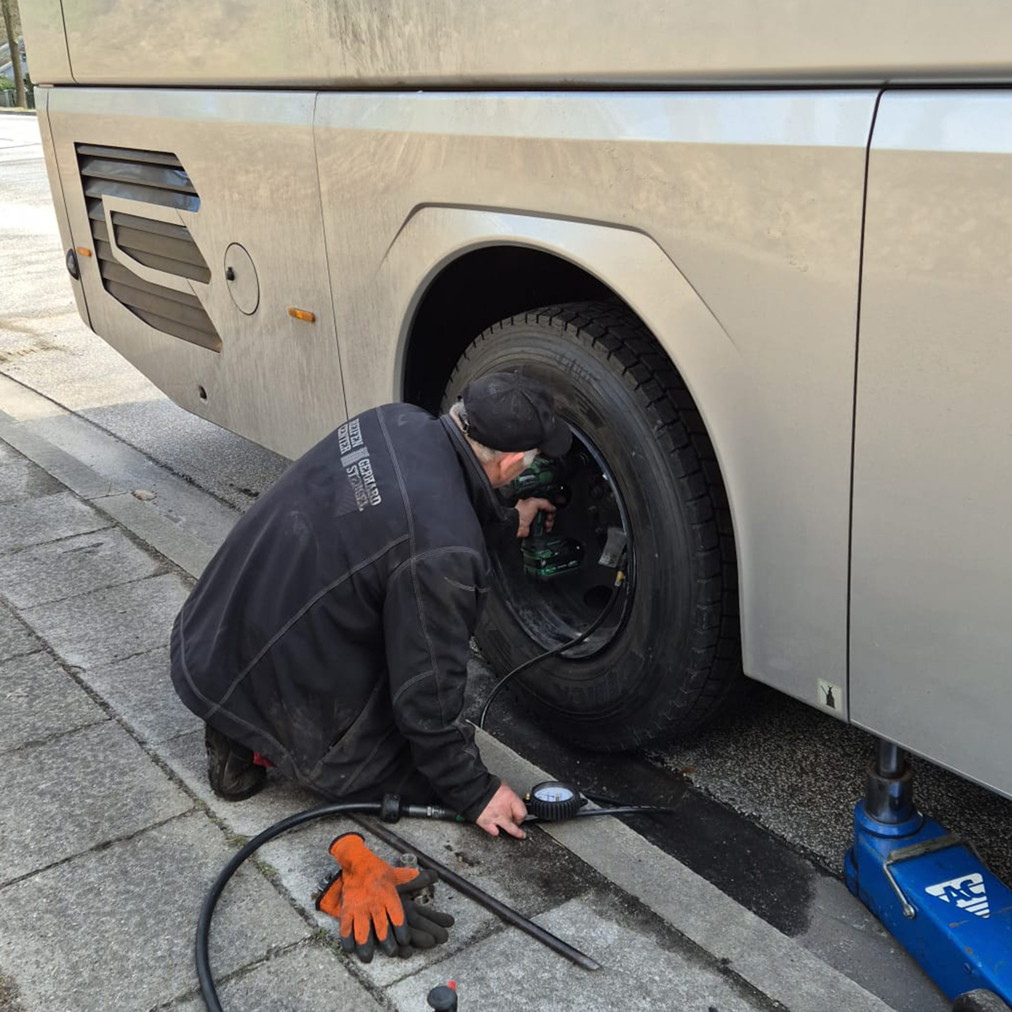 Eine Person arbeitet am Rad eines Busses, der auf einem Parkplatz steht. Werkzeuge und Materialien liegen auf dem Boden neben dem Bus.